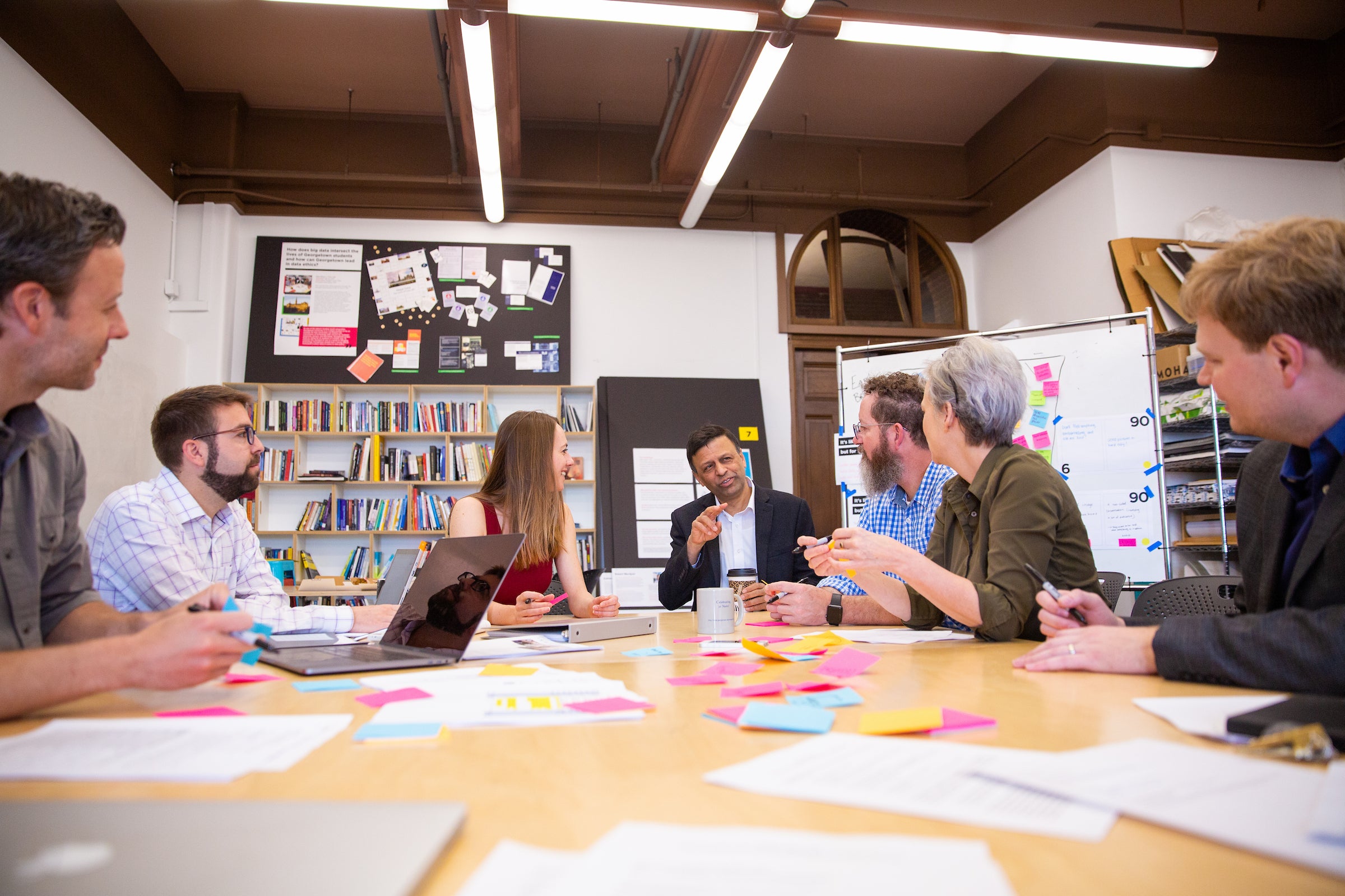 A group of people seated around a table, in conversation. The table is covered in paper and post-it notes. A whiteboard with notes is behind them.