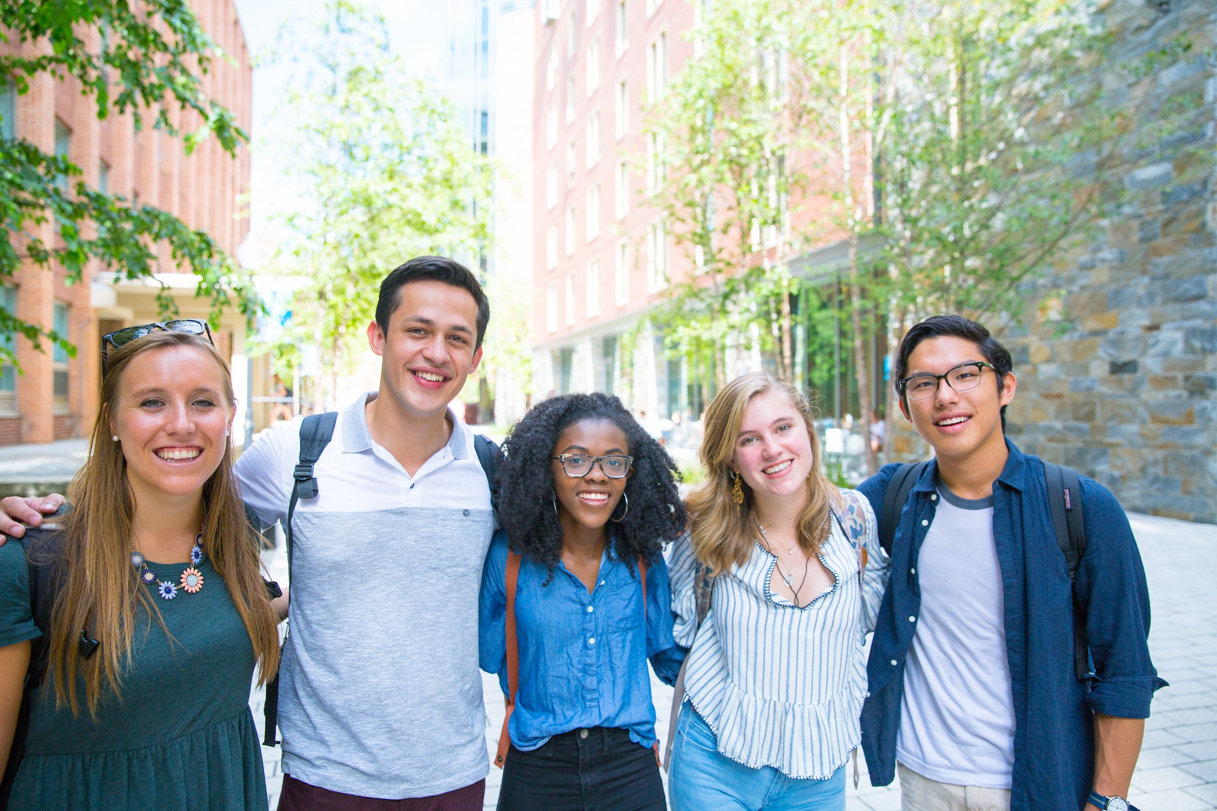 A group of 5 undergraduate students of mixed gender and race pose together outside, between two brick campus buildings.