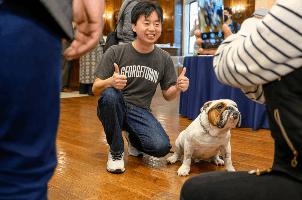 An Asian student wearing a Georgetown t-shirt and jeans, kneels beside Jack the Bulldog on a hard wood floor and gives 2 thumbs up.