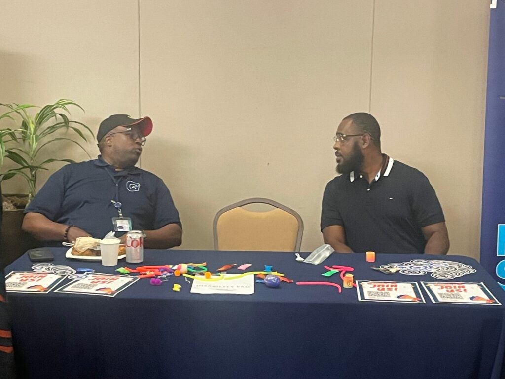 Two people sit, engaged in conversation from behind a table filled with flyers and giveaways.